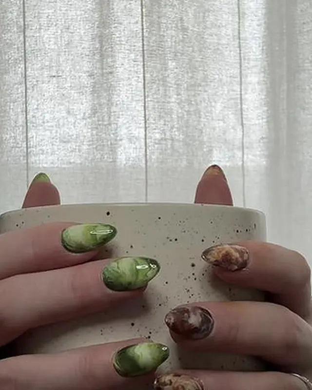Deux mains autour d’une tasse : ongles marbrés vert d’un côté, brun ambré de l’autre.